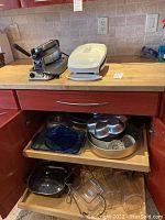 Photo of kitchen counter showing Waring Pro waffle maker stainless steel unit with control knob and light, white George Foreman electric grill, and open cabinet below displaying assorted bakeware including metal pans and glass bake dishes.