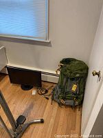 Photo showing Samsung monitor and green Habitat hiking backpack placed on wooden floor next to each other.