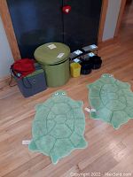 Full view of two green turtle rugs on floor with storage containers against wall including green cylindrical bin, gray foldable storage container and small plastic bins.