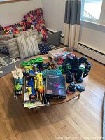 Overview of a collection of toys on a round wooden table including Nerf blasters, green remote cars, boxed Laser Pegs, and a black robot figure.
