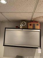 Front view of rectangular wall cabinet with white roller door and black frame. Key lock visible at top center.