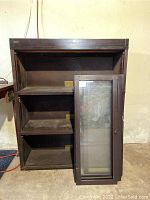 Front view showing the bookcase with one glass door resting against it revealing three shelves inside.
