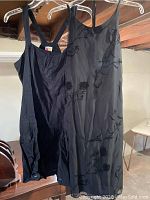 Photo of two black dresses hanging on hangers and displayed side by side on a rack against a basement wall and ceiling.