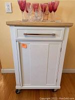 Front view of white wooden kitchen cart with butcher block top, single drawer and cabinet below, and wheels visible.