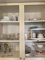 View of upper and middle shelves showing Corelle dishes with floral pattern, clear glass tumblers, wine glasses, white ceramic kitchen items, and rotating plastic cabinet organizer with multiple levels.