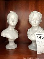 Pair of white plaster busts depicting boy and girl shown on wooden shelf.