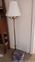 Full view of vintage brass floor lamp with white fabric lampshade and Holmes fan box at base.