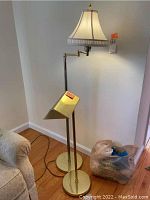 Two brass or gold-tone metal floor lamps of different heights side by side, one with adjustable arm and scalloped lampshade, other with fixed stand and pineapple finial on the lampshade.