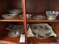 Various glass and silverplate serving dishes including divided serving plate, bowls and trivet on wood shelf inside cabinet.