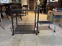 Black metal rolling cart front view with two shelves and caster wheels, step stool beside it.