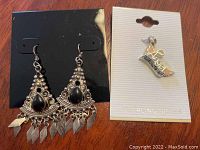 Photo of a pair of silver earrings with black teardrop stones and an ornate design, along with a sterling silver 'Love' charm on white card.