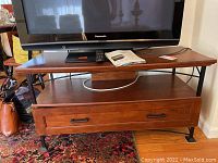 Full front view of wood and metal TV stand with drawer, showing top surface with TV and remote, drawer front with handles.
