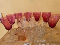 Photo showing seven to eight tall pink swirl pattern stemmed glasses, two clear wine glasses, two clear shot glasses, small glass pourer, and a clear glass basket with chip.