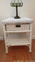 Front view of the white wicker bedside table showing drawer and bottom shelf with a lamp on top.