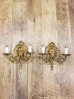 Pair of heavy solid brass Victorian wall sconces resting on wooden floor, showing full front view with two arms each for candelabra bulbs and push button switches visible