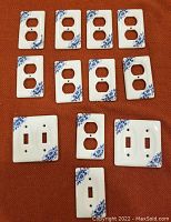 Top view of ten porcelain switch and outlet plates laid out on a reddish surface, showing the blue Delft style floral designs on white porcelain.