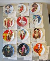 Photo of 12 Marilyn Monroe collectible plates laid out in four rows of three, each portraying a famous movie role of Marilyn Monroe, with certificates of authenticity beneath each plate.