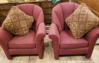 Front view of two burgundy upholstered chairs with one decorative pillow each, showing cushioned fan backs and arm protectors.