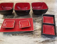 7 piece sushi set including 3 large square bowls, rectangular tray with 2 small square sauce dishes, 2 rectangular sauce dishes in a holder. Black exterior, red glossy interior.