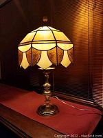 Vintage Tiffany style stained glass lamp illuminated, showing amber, beige, taupe, and cream glass colors with scalloped pattern and brass base.