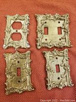Four solid heavy brass wall plates with ornate vintage floral and scroll design on red surface. Plates include single toggle, double toggle, and duplex outlet. Shows scratches and crazing.