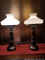 Pair of vintage Victorian style parlor lamps with white milk glass shades and carved wooden bases. Lamps are shown from front view with light on, highlighting the milk glass shades and brass holders.