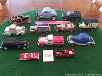 Photo showing all 12 die cast model vehicles arranged on a table with a green mat underneath, displaying various vintage style cars and trucks.