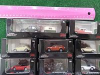 14 die-cast international toy cars in original black packaging with clear windows, laid out on green surface with pink ruler for scale.