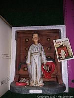 Front view of the figurine placed in protective white foam, showing the boy in pajamas and the dresser behind him, holding a red bag with a small animal.