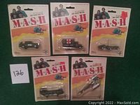 Five original 1976 M*A*S*H die cast metal toys displayed in original sealed packaging. Includes a military jeep, ambulance, staff car, transport truck, and helicopter. Pink ruler shown for scale.
