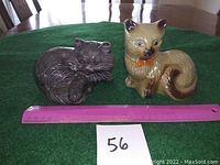 Photo shows both ceramic cat figurines side by side on a green surface with a ruler for size reference.