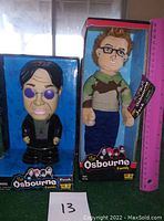 Front view of Ozzy Osbourne bank in blue themed box and Jack Osbourne plush doll in red and black themed box, next to a pink ruler for scale.