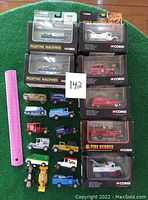 Photo of twenty CORGI die cast toy vehicles including boxed Fighting Machines tanks, boxed Fire Heroes emergency vehicles, and various loose miniature cars and trucks against a green background with a pink ruler for scale.
