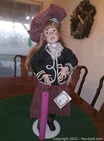 Front view of the Scottish porcelain doll showing detailed facial features, red tartan skirt, black jacket, and matching beret with green tassel. Doll pinned to white circular stand; purple ruler is placed in front for scale.