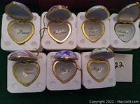 Seven heart-shaped enamel lockets shown open with inscriptions inside on a white protective foam base.