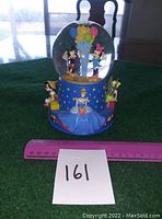 Full front view of the Disney musical snow globe showing Mickey and Minnie inside the globe and surrounding characters on the blue base. A pink ruler for size reference and lot card '161' in front.