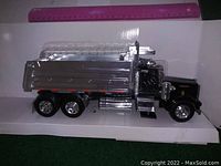 Side view of large silver toy dump truck showing six wheels, cab with roof details, and shiny dump bed in white cardboard box with ruler for scale.