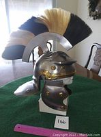 Side view of large metal helmet with black and blonde mohawk crest on mannequin head stand. Shows brass rivet detailing and cheek guards.