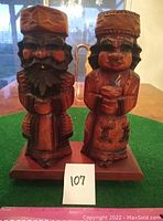 Front view of the two carved wooden figures on a wood base, showing details on faces and attire, along with lot number 107.