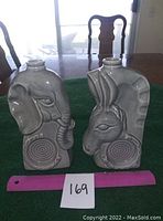 Front view showing grey ceramic Elephant and Donkey head decanters on green surface with pink ruler and lot number.