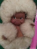 Front view close-up of the doll's head and upper fur parka hood, showing molded dark brown face with playful expression and fuzzy beige parka.