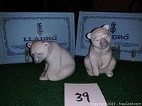 Two white porcelain Lladro polar bear figurines on green surface with their original branded boxes behind them, showing model details