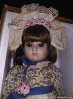 Close-up of doll face showing porcelain face, blue eyes, brown hair, floral dress with lace bonnet and ribbon flowers.