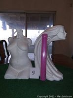 Two white statues showing female forms, next to pink rulers for scale, placed on a table indoors with a window background.