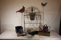 Full lot arranged on a white surface showing folding iron plant holder with three flowerpots, two iron bird garden stakes, decorative metal wall hanging, watering can, small glass pots, wooden flowerpot holder, and garden accessories.