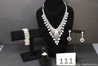 Photo showing entire jewelry set displayed on black stands, including necklace on bust, bracelet on bar, earrings on stand, and ring, with lot number sign 111 in foreground.