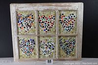 Front view of old wooden window frame containing six mosaic heart panes with different colored glass pieces on a green and yellow tiled background.