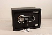 Front view of closed black rectangular steel safe with keypad, fingerprint sensor, handle, and keys inserted.