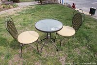 Full view of the round metal frame patio table with glass top and two metal chairs with fabric cushions, shown outdoor on lawn.