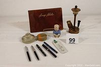 All seven items arranged showing porcelain man, lamp piece, glass paperweight, fountain pens, mechanical pencil, ribbon box, and souvenir book.